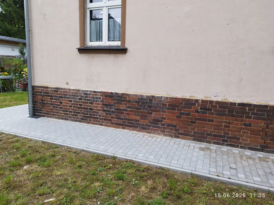 Verputzte Hauswand mit dunklem Klinkersockel und gepflastertem Weg
